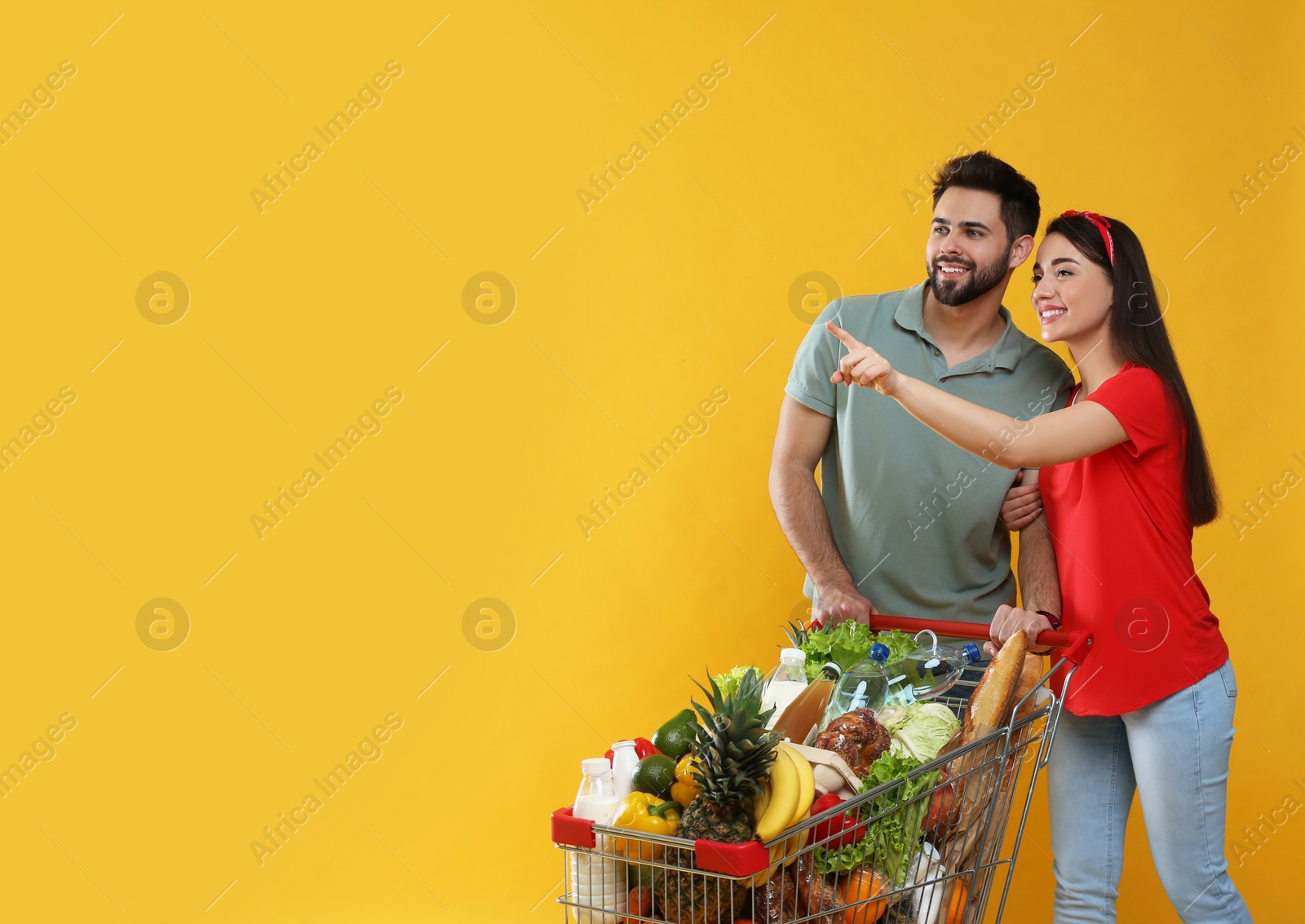 Photo of Young couple with shopping cart full of groceries on yellow background