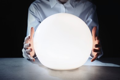 Photo of Businesswoman using glowing crystal ball to predict future at table in darkness, closeup. Fortune telling
