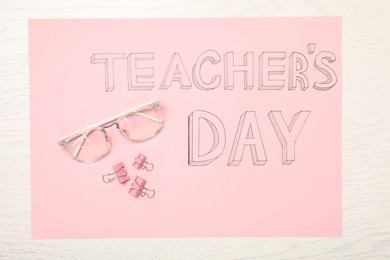 Sheet of pink paper with words TEACHER'S DAY, glasses and clips on white wooden background, top view