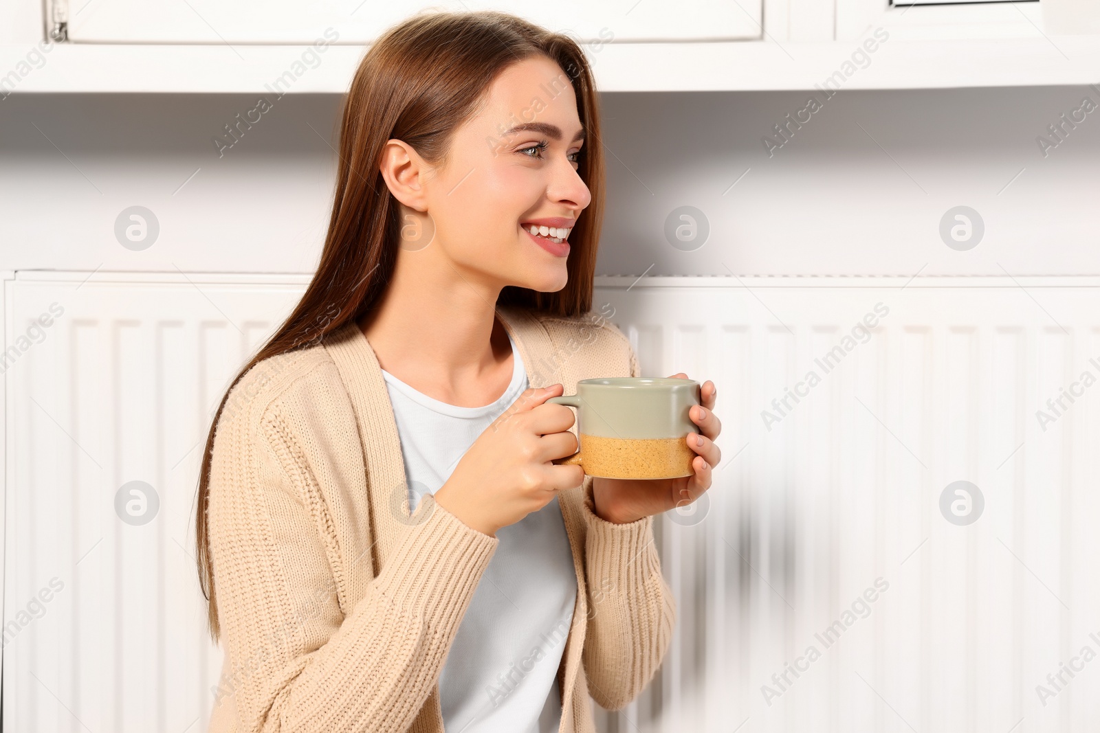 Photo of Woman holding cup with hot drink near heating radiator indoors