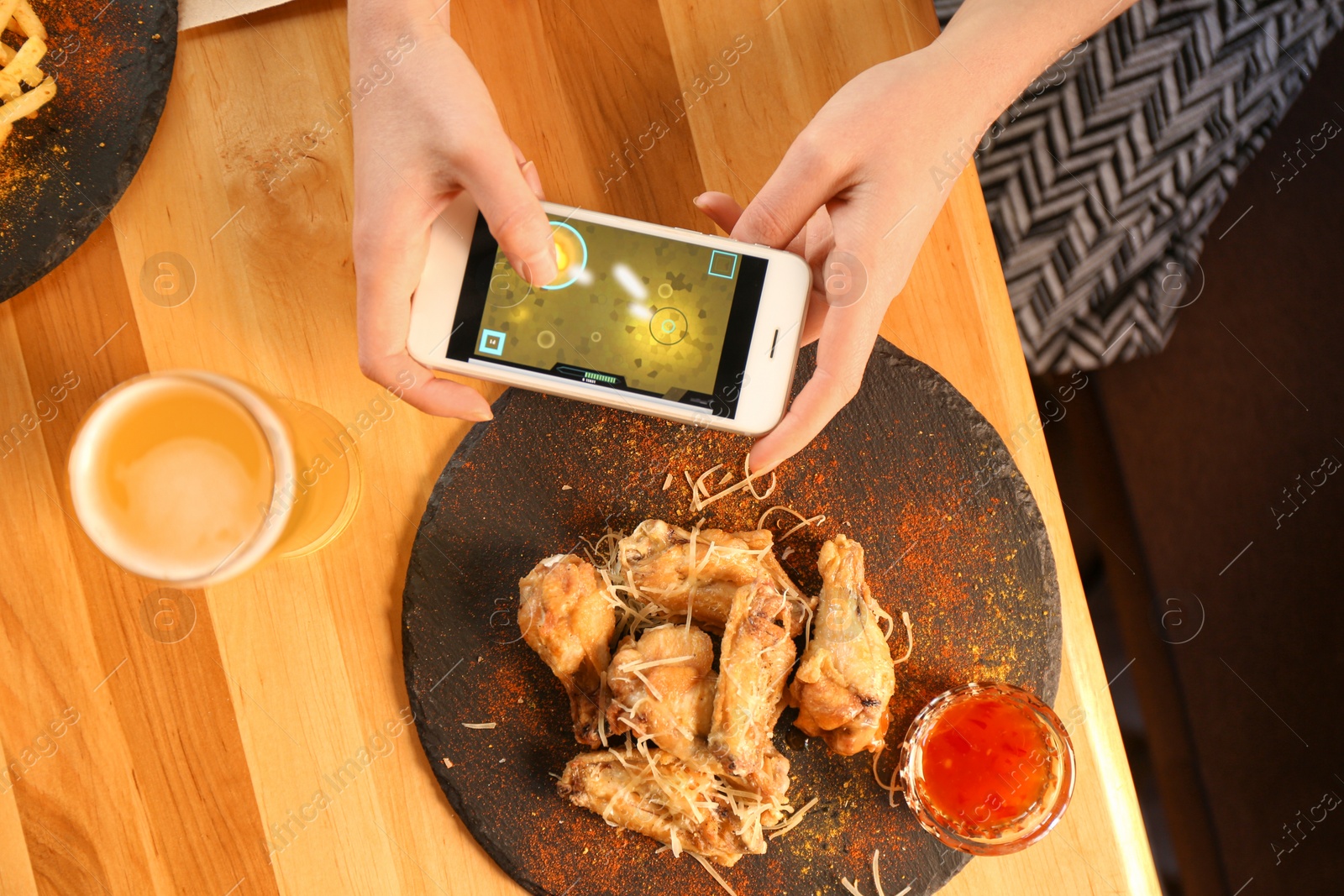 Photo of Woman playing game using smartphone at table with tasty BBQ wings, top view