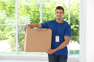 Happy young courier with cardboard box and clipboard near window indoors