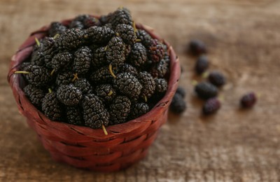 Wicker basket with delicious ripe black mulberries on wooden table, space for text