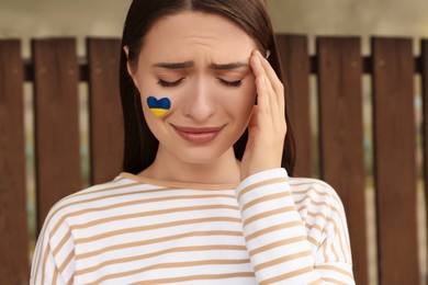 Sad young woman with drawing of Ukrainian flag on face outdoors
