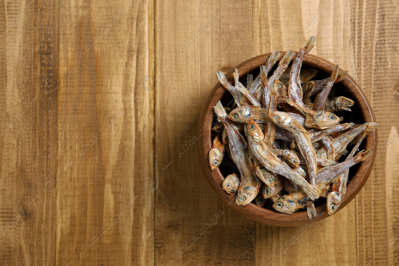 Photo of Bowl of tasty dried anchovies on wooden table, top view. Space for text