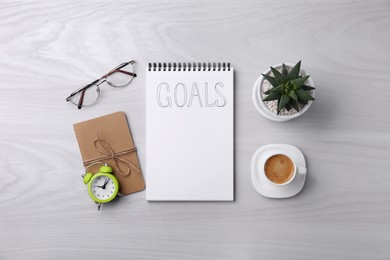 Notebook with word Goals, houseplant, alarm clock, glasses and cup of coffee on white wooden table, flat lay. Planning concept