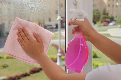 Photo of Young woman cleaning window glass with rag and detergent at home, closeup