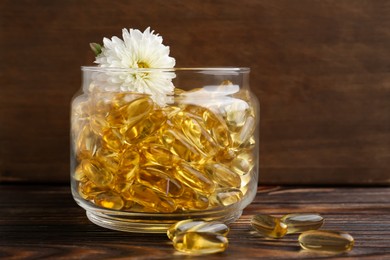 Photo of Glass medical bottle with yellow capsules and white flower on wooden table, closeup