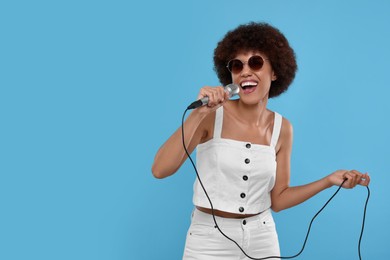 Curly young woman with microphone singing on light blue background, space for text