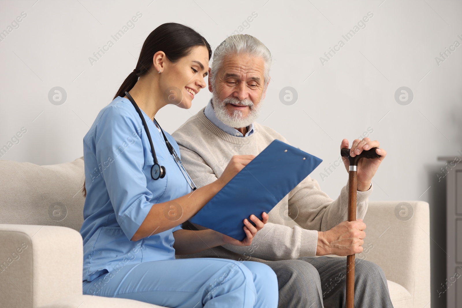 Photo of Smiling nurse with clipboard assisting elderly patient on sofa in hospital