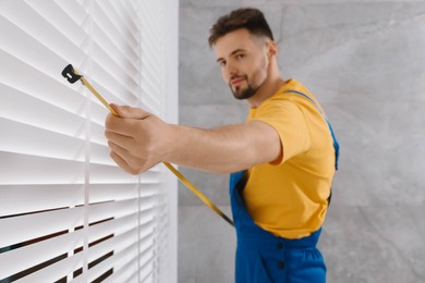 Worker in uniform using measuring tape while installing horizontal window blinds indoors, focus on hand