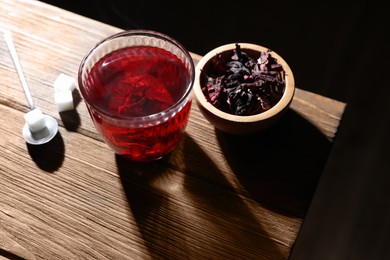 Photo of Delicious hibiscus tea in glass, sugar cubes and dry roselle petals on wooden table