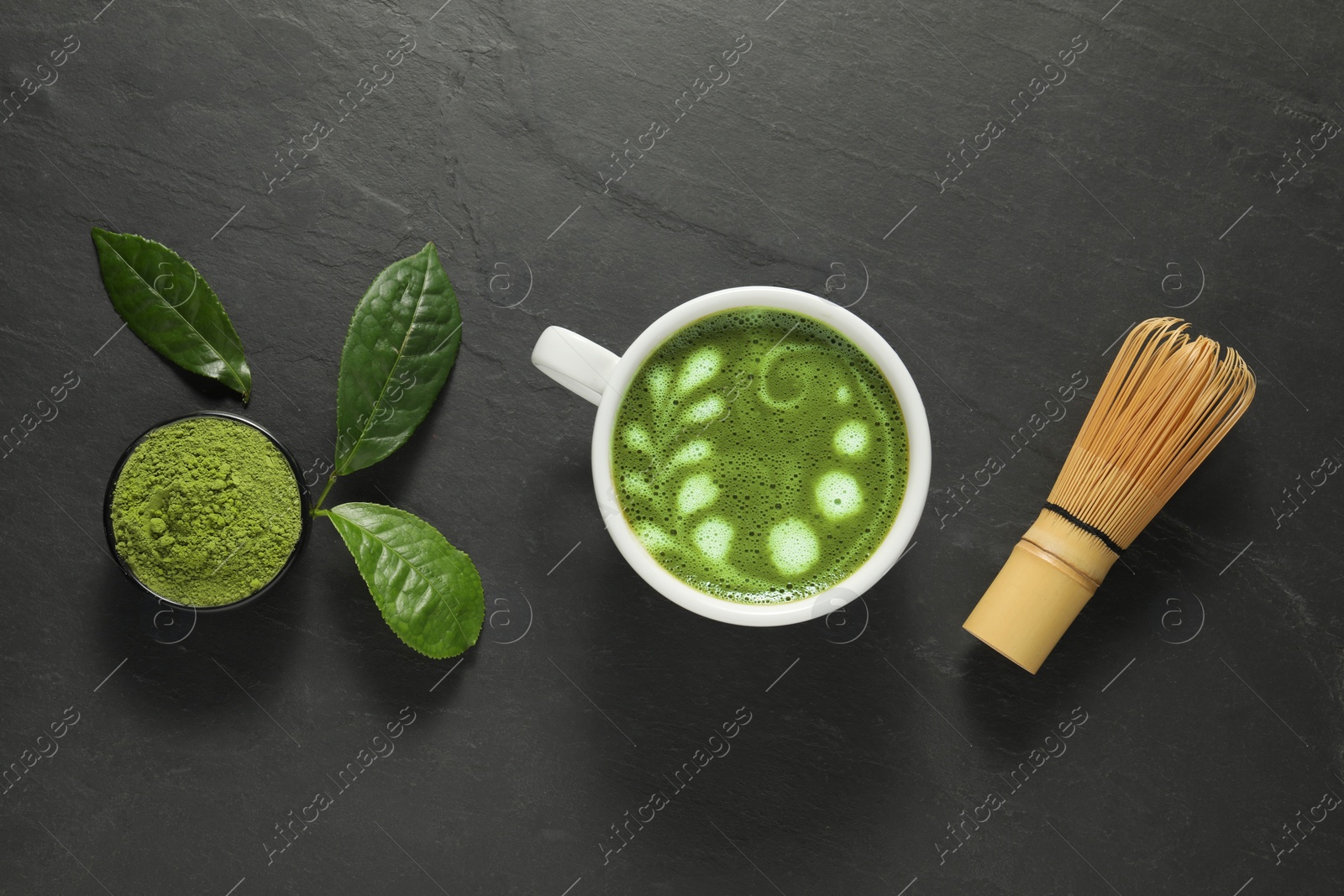Photo of Delicious matcha latte, powder, leaves and whisk on dark textured table, flat lay