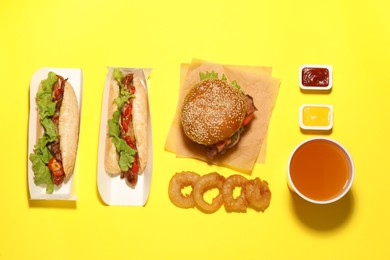 Photo of Tasty burger, hot dogs, fried onion rings, different sauces and refreshing drink on yellow background, flat lay. Fast food