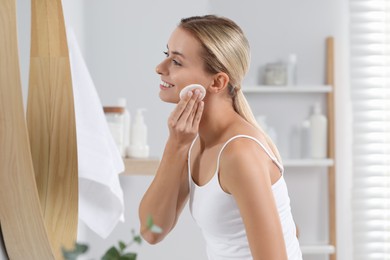 Smiling woman removing makeup with cotton pad in front of mirror in bathroom