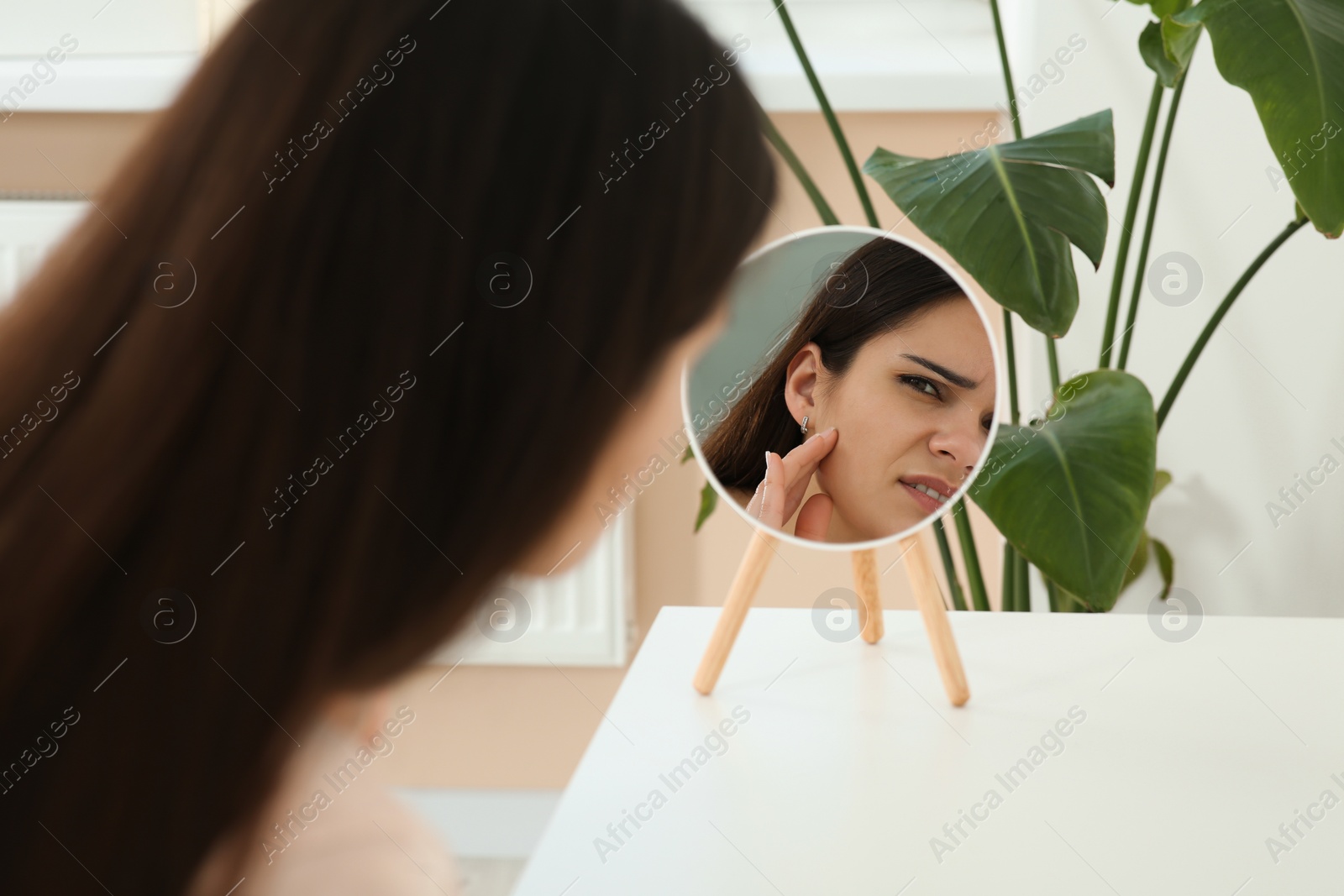 Photo of Young woman looking in mirror and touching her face indoors. Hormonal disorders