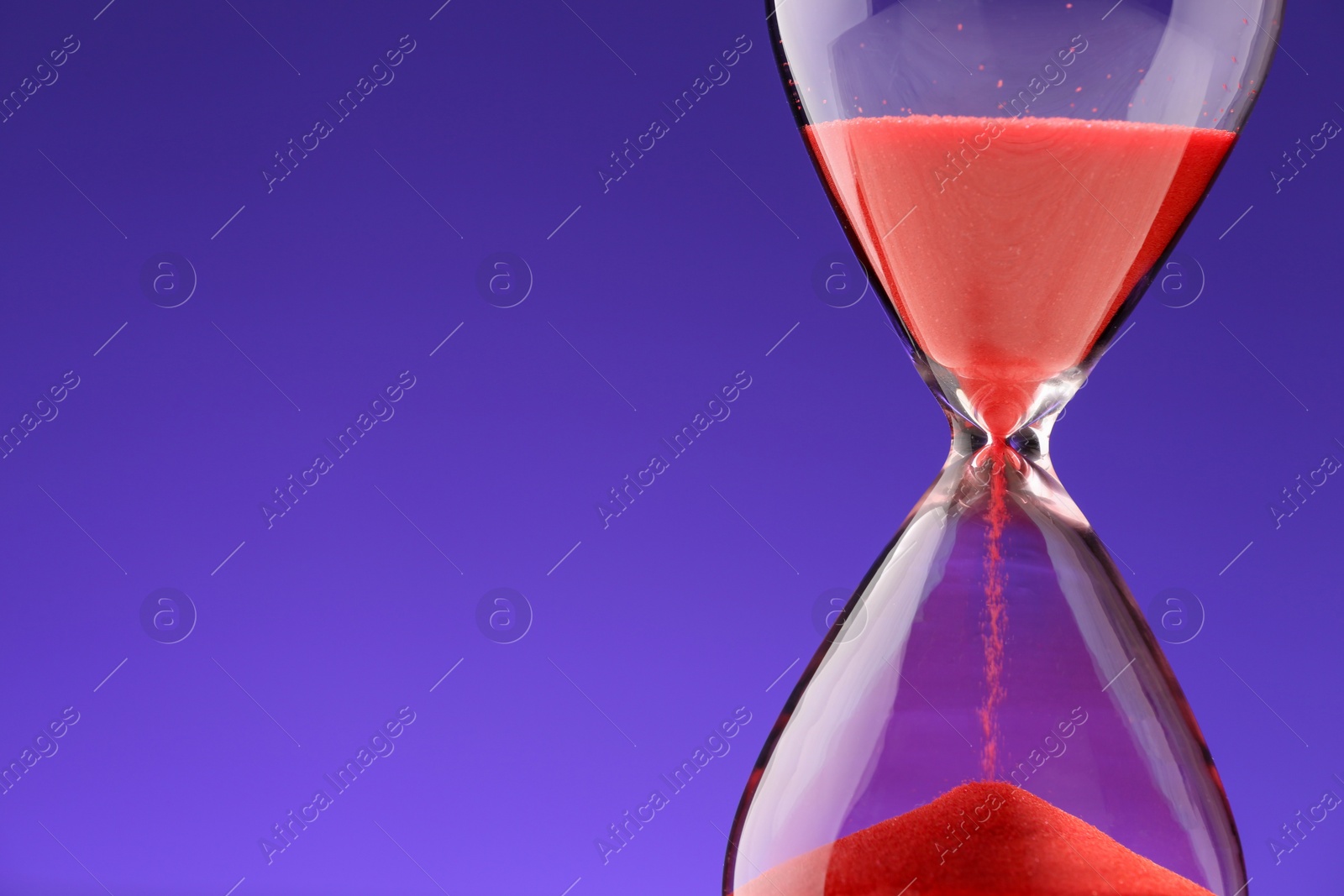 Photo of Hourglass with red flowing sand on purple background, closeup. Space for text