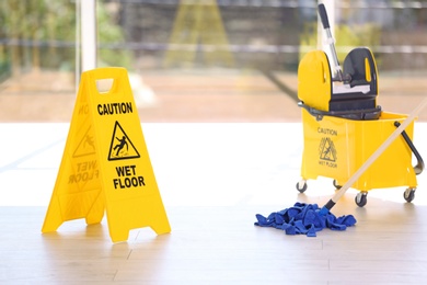 Photo of Safety sign with phrase Caution wet floor and mop bucket, indoors. Cleaning service