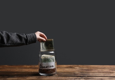 Woman putting money into donation jar on wooden table against grey background, closeup. Space for text