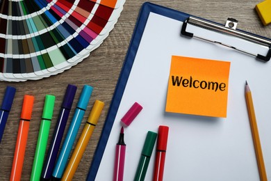 Image of Sticky note with word Welcome, clipboard, colorful markers and palette on wooden table, flat lay