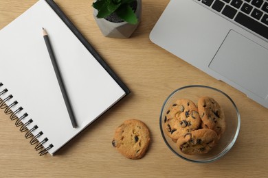 Photo of Chocolate chip cookies, notebook and laptop on wooden table at workplace, flat lay