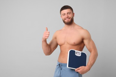 Photo of Happy athletic man holding scales and showing thumb up on light grey background, space for text. Weight loss concept