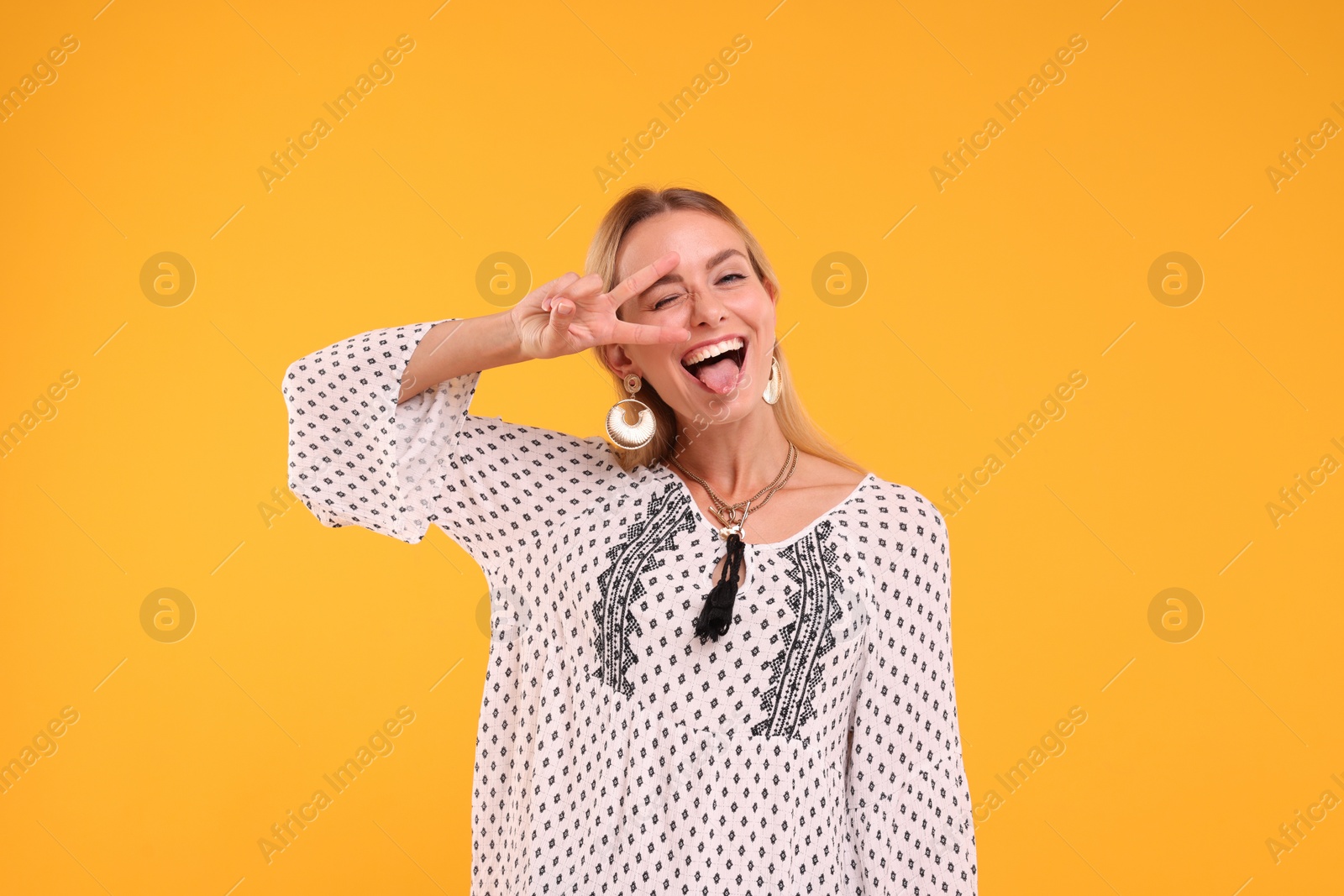 Photo of Portrait of hippie woman showing tongue and peace sign on yellow background