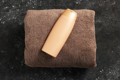 Photo of Soft brown terry towel and cosmetic bottle on black textured background, top view