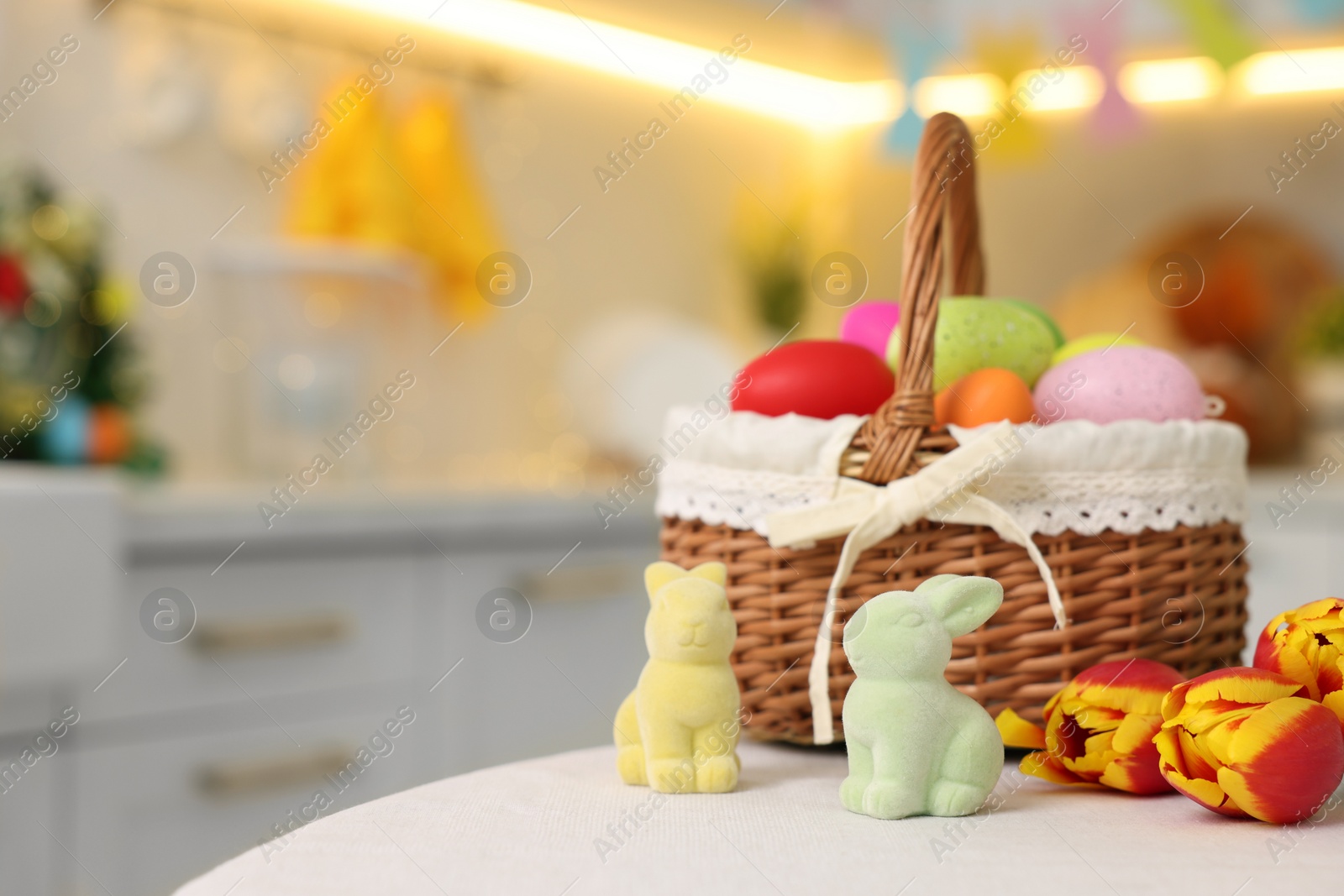 Photo of Easter decorations. Wicker basket with painted eggs, tulips and bunny figures on white table indoors, closeup. Space for text