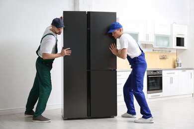 Professional workers carrying modern refrigerator in kitchen