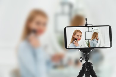 Beautiful woman applying makeup near mirror in room, selective focus on smartphone display