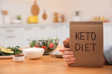 Woman holding notebook with phrase Keto Diet and different products at wooden table in kitchen, closeup