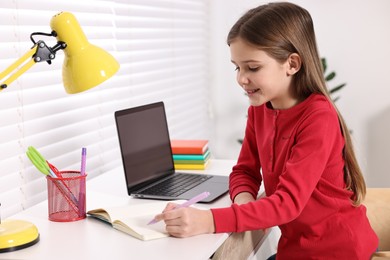 Photo of E-learning. Cute girl taking notes during online lesson at table indoors