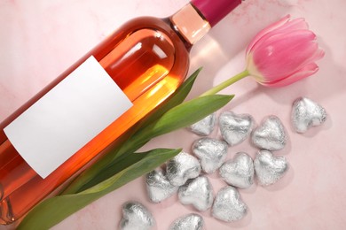 Delicious heart shaped chocolate candies, beautiful tulip and bottle of wine on pink table, flat lay