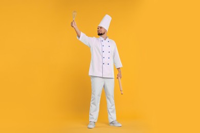 Photo of Happy professional confectioner in uniform holding rolling pin and whisk on yellow background