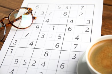 Sudoku, cup of coffee and eyeglasses on wooden table, closeup