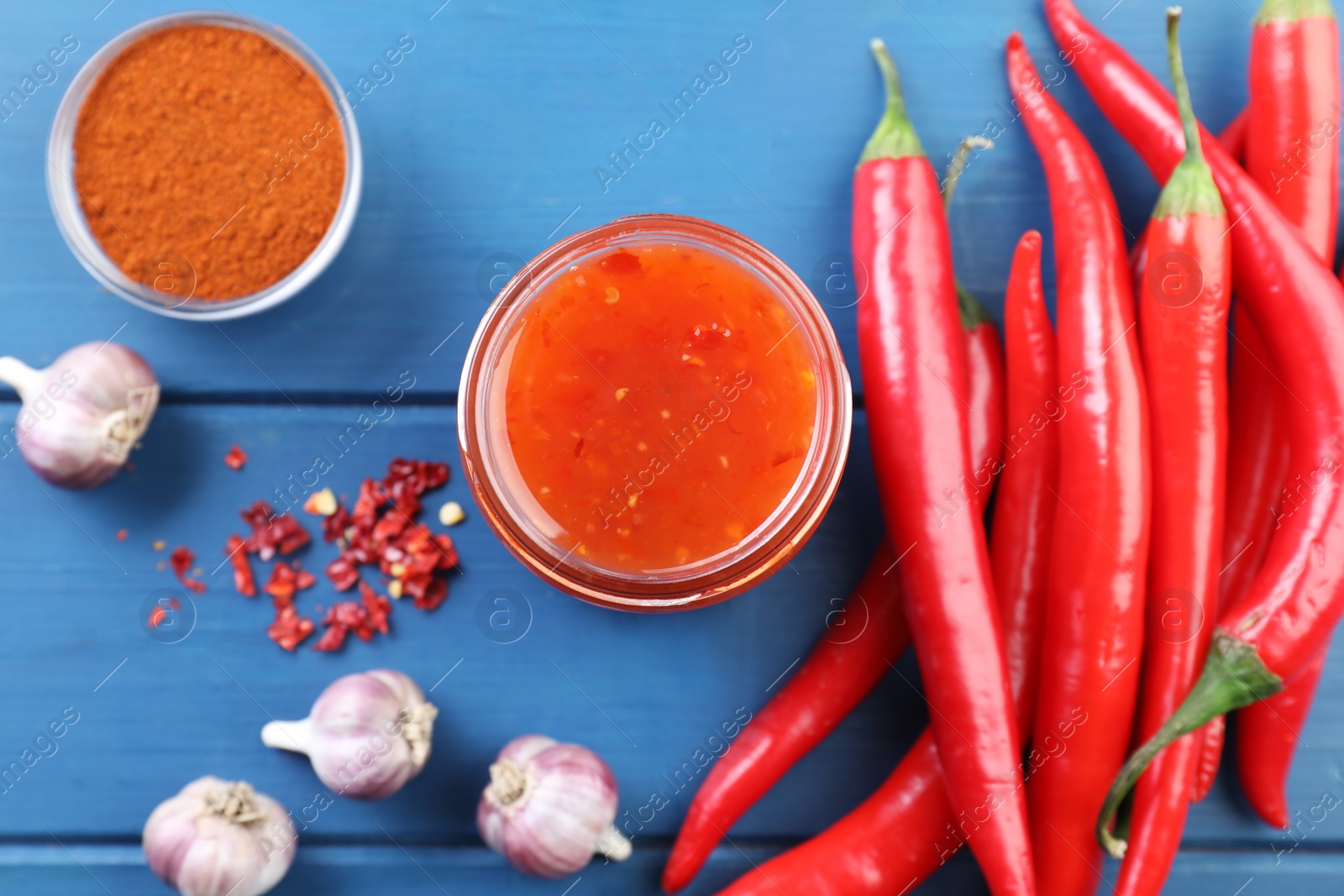 Photo of Spicy chili sauce in jar and ingredients on blue wooden table, flat lay