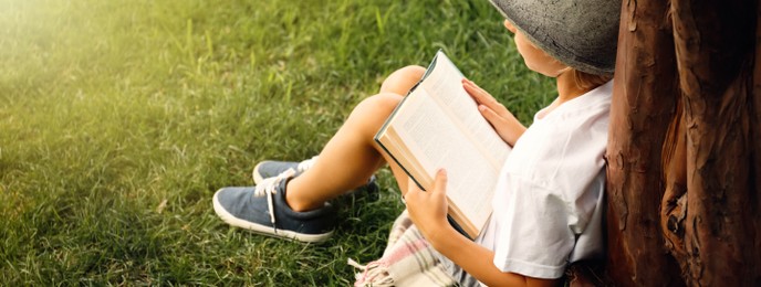 Image of Cute little boy reading book on green grass near tree in park, space for text. Banner design