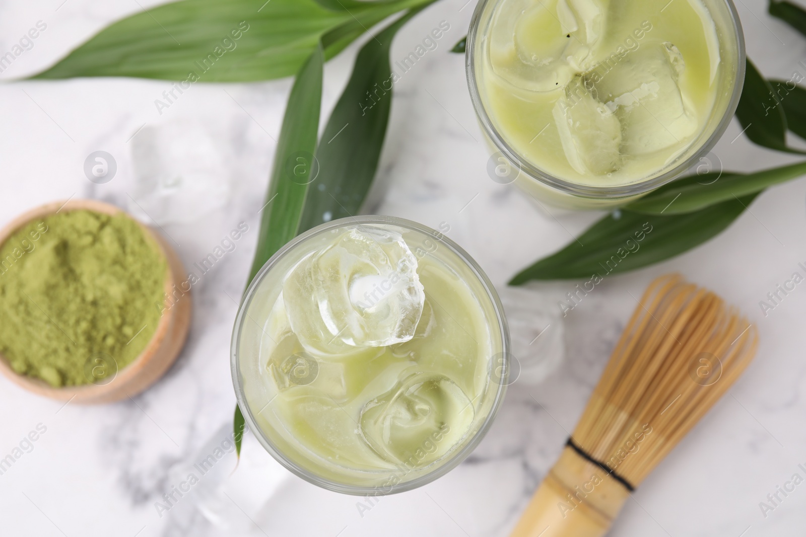 Photo of Glasses of tasty iced matcha latte, bamboo whisk and leaves on white marble table, flat lay