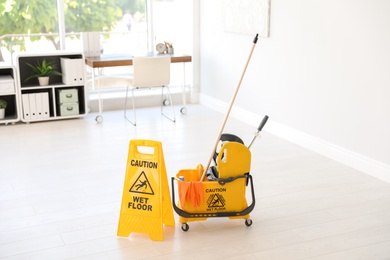 Photo of Phrase "CAUTION WET FLOOR" on safety sign and mop bucket with cleaning supplies, indoors