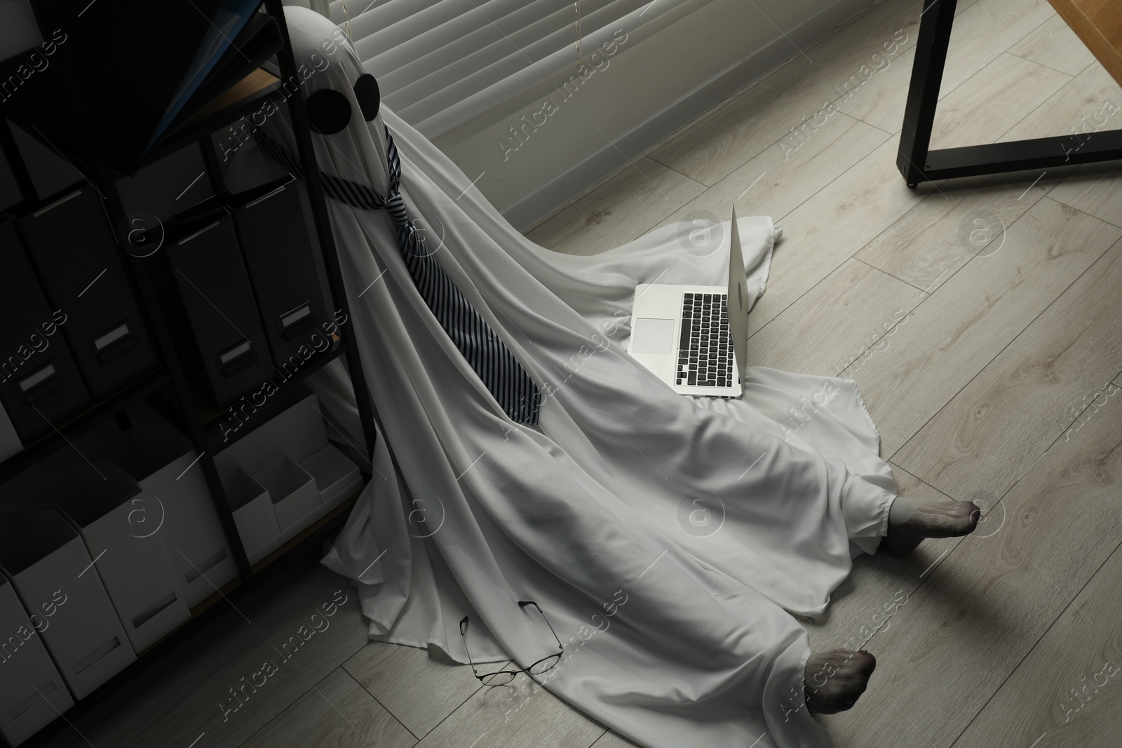 Photo of Overworked ghost. Man in white sheet with laptop on floor in office, above view