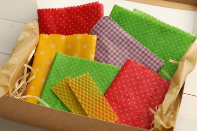 Colorful beeswax food wraps in box on white wooden table, closeup