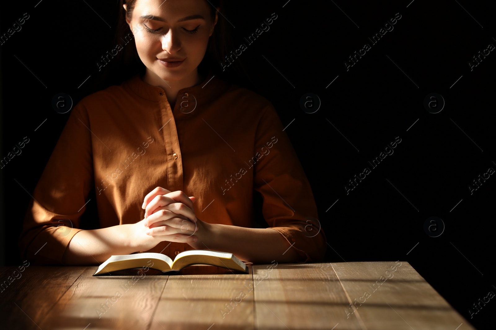 Photo of Religious young woman praying over Bible at wooden table indoors