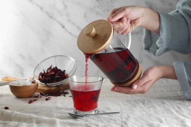 Woman pouring delicious hibiscus tea from glass teapot into cup at table, closeup