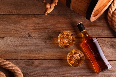 Photo of Whiskey with ice cubes in glasses, bottle and barrel on wooden table, flat lay. Space for text