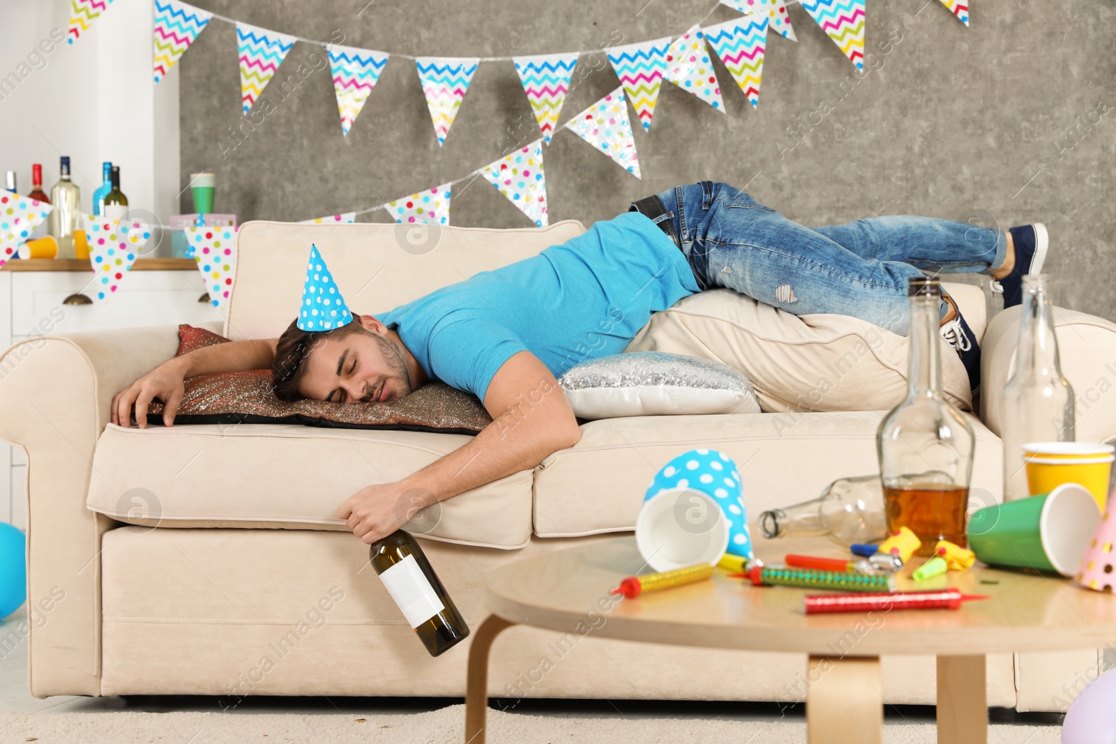 Photo of Young man with bottle of wine sleeping on sofa in messy room after party