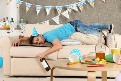 Young man with bottle of wine sleeping on sofa in messy room after party