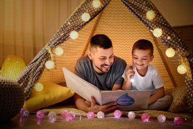Father and son with flashlight reading book in play tent