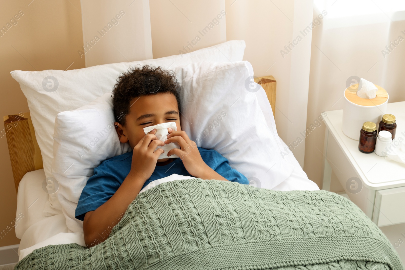 Photo of African-American boy with scarf and tissue blowing nose in bed indoors, above view. Cold symptoms