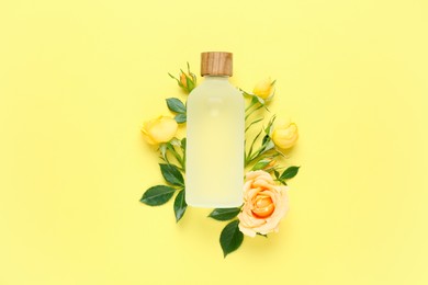 Bottle of rose essential oil and beautiful flowers on pale yellow background, top view